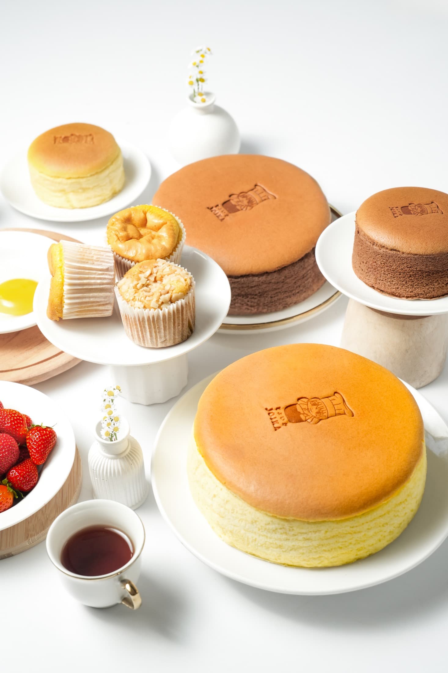 Rangkaian Japanese cheesecake original, cokelat, ukuran personal, dan Bolu Joika di atas meja putih.